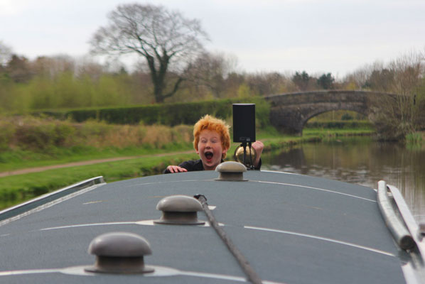 a child looks delighted as he stands at the helm of a narrowboat behind him is a bare tree and a stone bridge
